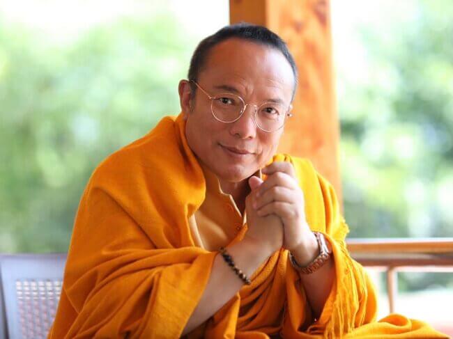 Se aprecia a Tai Situ Rinpoche en primer plano. Se ve como se toma sus dos manos de forma relajada. A su espalda se ve un lugar verde y una viga de madera.